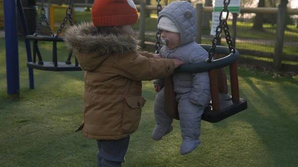 Komik, küçük, tombul kız, ablası, yeni doğmuş erkek kardeşi salıncakta sallıyor. Bebek oyun parkında oynuyor. Çocuk Eğlencesi, Çocukluk, Çocuk Geliştirme, Mutlu Aile Kavramı — Stok fotoğraf