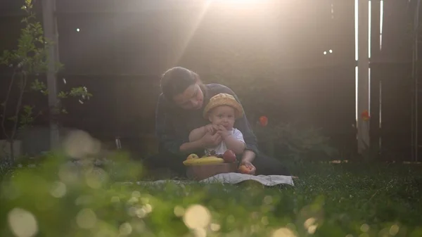 Boldog fiatal, vidám anya tartja baba eszik gyümölcsöt a zöld fű. Anya imádnivaló csecsemő gyermek játszik a szabadban szeretettel a kertben. Kisgyerek a szülőkkel. Család, természet, ökológia koncepció — Stock Fotó