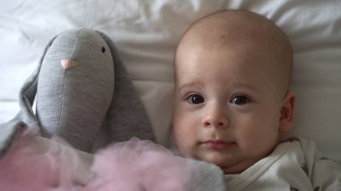 Yakın plan Mutlu neşeli çocuk 6 aylık. Yeni doğan çocuk, beyaz yumuşak yatakta banyo sonrası kameraya bakıyor. Bebek oyuncakla yatmadan önce uyandı. Çocukluk, annelik, aile, bebek konsepti