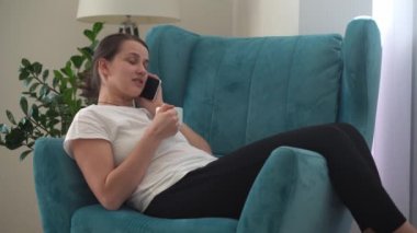 Oturma odasında telefonla konuşan otantik bir genç kadın. Kullanarak Yazma Araması. Smartphone 'da çalışan mutlu bayan rahat rahat koltukta internette geziniyor. Buisenes, Eğitim, Teknoloji Konsepti