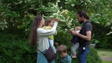 Happy family mother Father four Baby little siblings kids have fun sniffing flowers on trees enjoying summer holidays in garden park. Smiling parents children spending leisure time together sunset