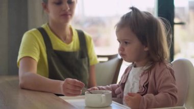 Yakından anne, beyaz giyen, yüksek sandalyedeki bebeği besliyor, çocuk kendini yemeye çalışıyor, yemek lekeli mutlu çocuk, küçük kız kaşıkla yulaf lapası yiyor. Mutfakta kaliteli kahvaltı.