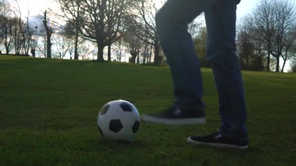 Человек в синих джинсах и мокасинах жонглирует классическим футбольным мячом. Люди занимаются спортом. Мальчик играет в футбол. Счастливая семейная команда провела время в парке. Здоровая жизнь, чемпионат — стоковое видео