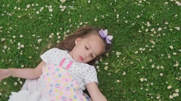 Malá šťastná holčička v šatech s lukem na hlavě ležící na zeleném trávníku v parku. Letní čas, příroda, sny, koncepty životního stylu. Usmívající se dětská tvář zblízka. emocionální dítě šklebí na kameru — Stock video