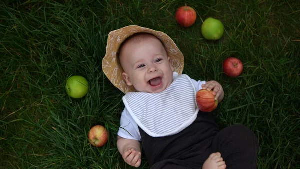 Happy Newborn child in summer panama hat Fall down laying on grass barefoot in Summer Sunny Day with Fresh Fruits Apples. Infant Kid Toddler Boy Smilling Face in Garden Healthy Food Nature harvest — стоковое фото