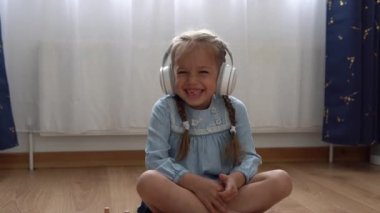 Büyük beyaz kulaklıkla müzik dinleyen çocuk. Anaokulundaki dişsiz kız 5 yaşında ve içerideki odada kameraya bakıyor. Gülümseyen Çocuk Evde Dil Gösteriyor. Çocukluk, Eğitim.