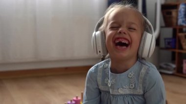 Büyük beyaz kulaklıkla müzik dinleyen çocuk. Anaokulundaki dişsiz kız 5 yaşında ve içerideki odada kameraya bakıyor. Gülümseyen Çocuk Evde Dil Gösteriyor. Çocukluk, Eğitim.