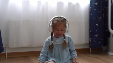 Büyük beyaz kulaklıkla müzik dinleyen çocuk. Anaokulundaki dişsiz kız 5 yaşında ve içerideki odada kameraya bakıyor. Gülümseyen Çocuk Evde Dil Gösteriyor. Çocukluk, Eğitim.