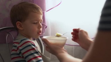Yemek, beslenme, çocukluk kavramları. Anne bebeği kaşık kaşık ile besliyor. Önlük giymiş şirin, aç bir çocuk çocuk çocuk sandalyesine oturur. Kapalı tabaktan yemek yemek istemez. Önce sevme