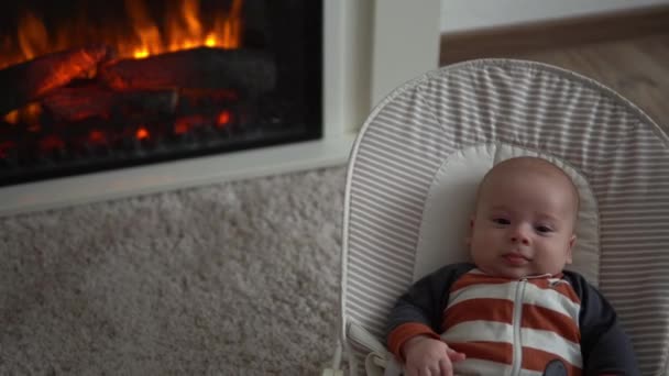赤ちゃんの様子をカメラの幼児 親の愛 ストライプのボディスーツを着たかわいい笑顔の新生児は 自宅で暖炉の近くの椅子の上に寝そべって見て目を覚ます 楽しい生活幸せな子供 — ストック動画