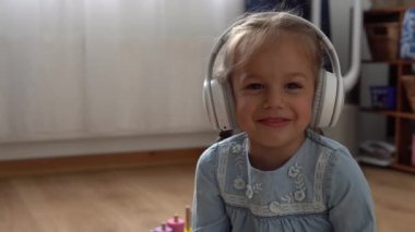 Büyük beyaz kulaklıkla müzik dinleyen çocuk. Anaokulundaki dişsiz kız 5 yaşında ve içerideki odada kameraya bakıyor. Gülümseyen Çocuk Evde Dil Gösteriyor. Çocukluk, Eğitim.