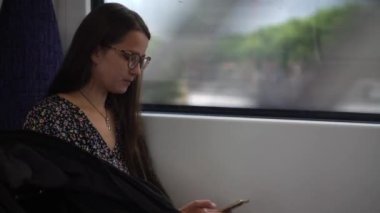 Close shot of young attractive Ukraine female in glasses sitting on moving city metro train. Woman calling typing text message on mobile phone. Pretty Lady waving hands say goodbye. Traveling alone.