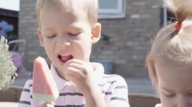 Close up portrait Boy enjoys delicious ice cream cone. Child eating watermelon popsicle. Kid Sibling snack sweets in Home Garden. Summer holiday Hot weather Sunny Day. Childhood, Food Candy Friends.