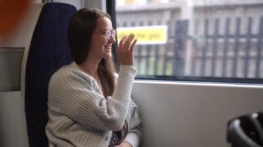 Close shot of young attractive Ukraine female in glasses sitting on moving city metro train. Woman calling typing text message on mobile phone. Pretty Lady waving hands say goodbye. Traveling alone.