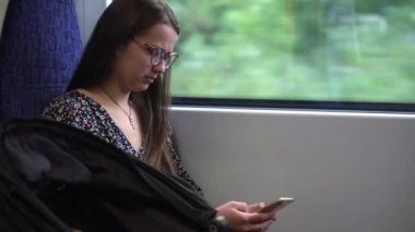 Close shot of young attractive Ukraine female in glasses sitting on moving city metro train. Woman calling typing text message on mobile phone. Pretty Lady waving hands say goodbye. Traveling alone.