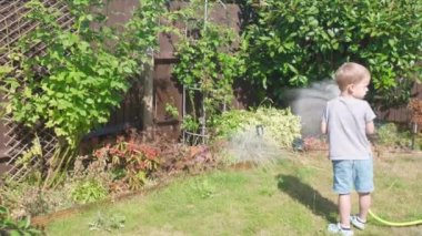 Bahçenin arka bahçesindeki çim bitkilerini sulayan komik küçük çocuk. Sıcak güneşli bir yaz gününde sulama hortumuyla oynayan sevimli bir çocuk. Çocuklar ev işlerine yardım eder. Çocuklar için etkinlik. Çocukluk.