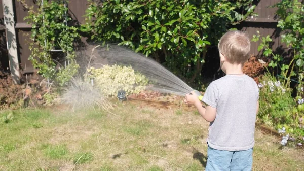 庭の住宅裏庭に面白い男の子の散水芝生の植物 暑い夏の屋外で灌漑ホースで遊ぶ愛らしい子供 子供は家事を手伝う 子供のための活動 幼少期 — ストック写真