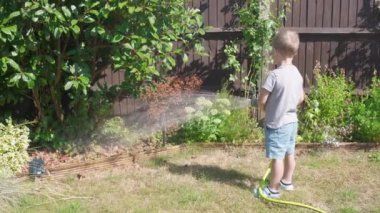 Bahçenin arka bahçesindeki çim bitkilerini sulayan komik küçük çocuk. Sıcak güneşli bir yaz gününde sulama hortumuyla oynayan sevimli bir çocuk. Çocuklar ev işlerine yardım eder. Çocuklar için etkinlik. Çocukluk.