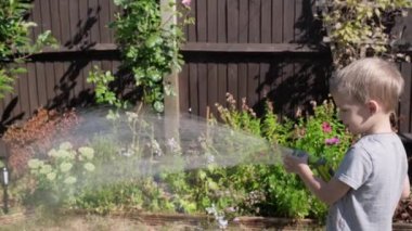 Bahçenin arka bahçesindeki çim bitkilerini sulayan komik küçük çocuk. Sıcak güneşli bir yaz gününde sulama hortumuyla oynayan sevimli bir çocuk. Çocuklar ev işlerine yardım eder. Çocuklar için etkinlik. Çocukluk.