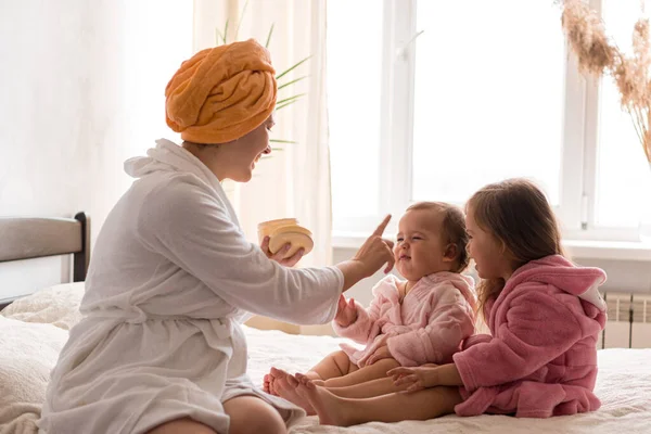 Young mother two daughters in curlers bathrobes. happy smiling family skin care concept. Mom teaches little kids child to do makeup smear cream on face sittting in orange bathrobes on bed in bedroom.