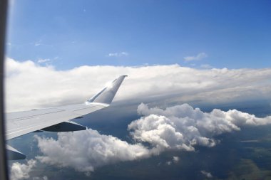 An aircraft wing flying in the blue sky and white clouds floating above green farmland on a sunny day. Viewed from an airplane window.