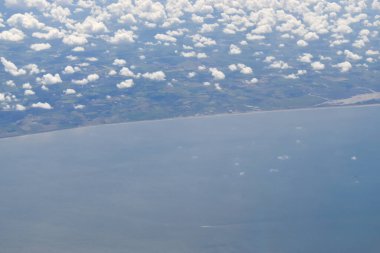 See-through from an aircraft window at a high angle with white clouds floating over a green landscape meeting with beaches and the sea, on a sunny day.