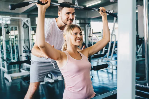Cheerful young woman wearing pink sports bra while doing chin-up exercise with trainer man in fitness gym