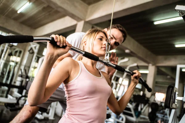 Cheerful young woman wearing pink sports bra while doing chin-up exercise with trainer man in fitness gym