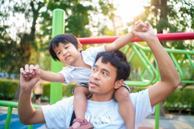 Küçük çocuk dışarıda oyun parkında eğlenirken babasının boynuna biniyor.
