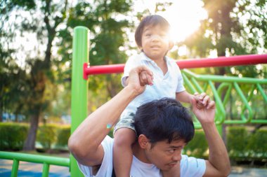 Küçük çocuk dışarıda oyun parkında eğlenirken babasının boynuna biniyor.