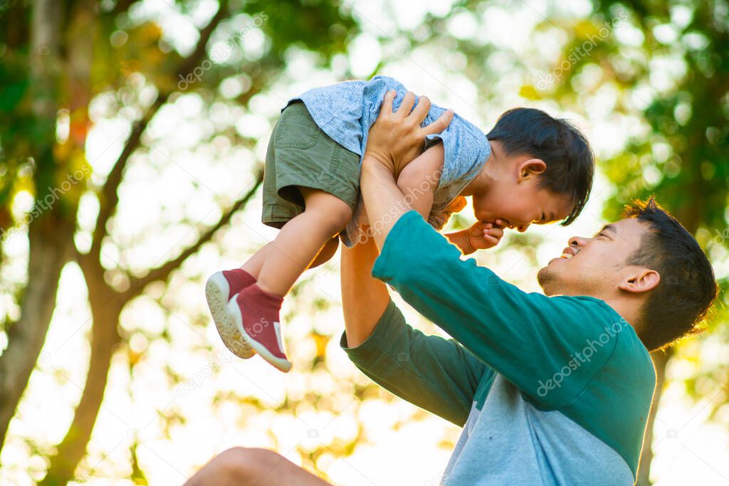 Father hold and lifting little boy in city park sunset light blurred green forest background