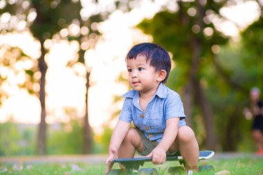 Küçük anaokulu çocuğu kaykay tahtasına oturmuş parkta çim üzerinde pratik yapıyor.