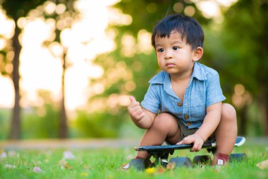 Küçük anaokulu çocuğu kaykay tahtasına oturmuş parkta çim üzerinde pratik yapıyor.