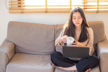 Bir dairede kanepede oturan bir kadın kahve içer ve ev işleri konseptinde çalışırken dizüstü bilgisayar açar..