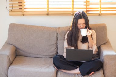 Bir dairede kanepede oturan bir kadın kahve içer ve ev işleri konseptinde çalışırken dizüstü bilgisayar açar..
