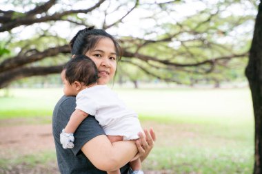 Güzel, genç Asyalı anne ağacın altında yeşil çimlerde oturuyor. Yanında da yaz çayırı yeşil çimen ağaçları var.