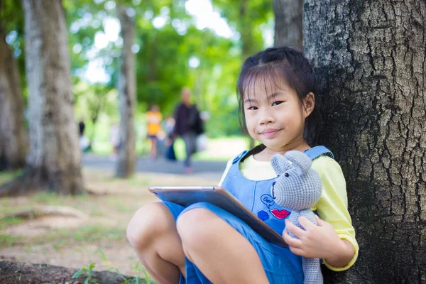 Ağaç parkında oturan sevimli küçük kız oyuncak bebekle tablet kullanıyor.