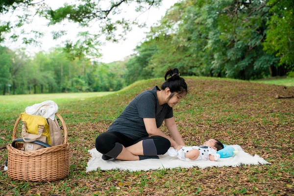 Güzel, genç Asyalı anne ağacın altında yeşil çimlerde oturuyor. Yanında da yaz çayırı yeşil çimen ağaçları var.
