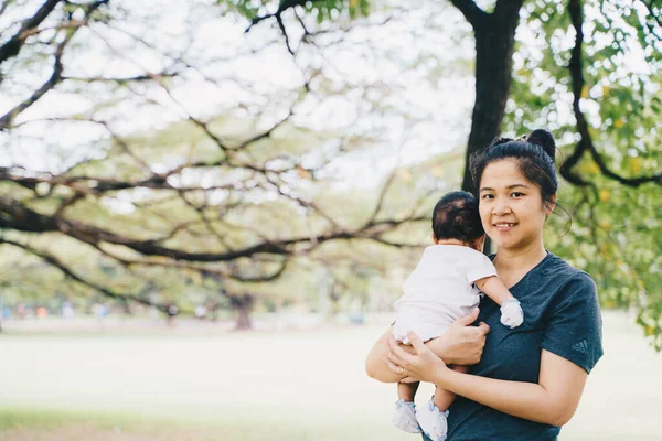 Güzel, genç Asyalı anne ağacın altında yeşil çimlerde oturuyor. Yanında da yaz çayırı yeşil çimen ağaçları var.