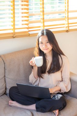 Bir dairede kanepede oturan bir kadın kahve içer ve ev işleri konseptinde çalışırken dizüstü bilgisayar açar..