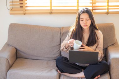 Bir dairede kanepede oturan bir kadın kahve içer ve ev işleri konseptinde çalışırken dizüstü bilgisayar açar..