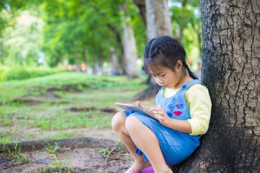 Ağaç parkında oturan sevimli küçük kız oyuncak bebekle tablet kullanıyor.