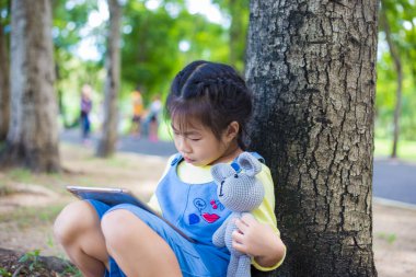 Ağaç parkında oturan sevimli küçük kız oyuncak bebekle tablet kullanıyor.