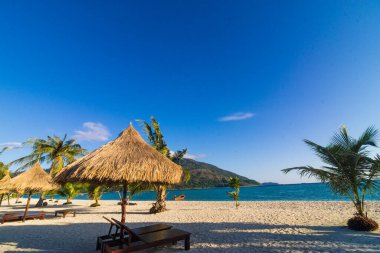 Tahta plaj sandalyeleri ve beyaz kumsalda kuru bitki şemsiyesi.