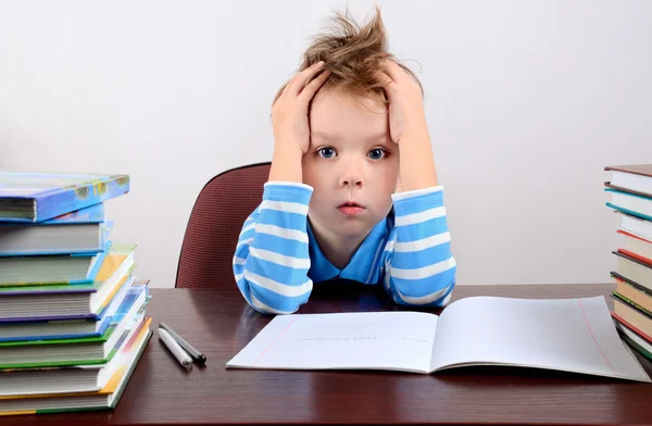 Lille træt dreng sidder ved et skrivebord og holder hænder mod hoved - Stock-foto