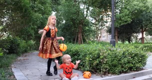 Cute little caucasian kids with blonde hair seven years old in costume of witch with hat and baby one year old celebrating Halloween outdoor on street. Holiday concept. Slow motion. — Stock Video