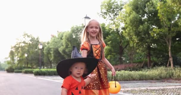 Roztomilé malé kavkazské děti s blond vlasy sedm let v kostýmu čarodějnice s kloboukem a dítě jeden rok slaví Halloween venku na ulici. Koncept dovolené. Zpomalený pohyb. — Stock video