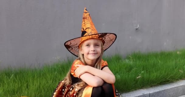 Linda niña caucásica con el pelo rubio de siete años de edad en traje de bruja con sombrero y vestido naranja negro celebrando Halloween al aire libre en la calle. Concepto de vacaciones. Movimiento lento. — Vídeo de stock