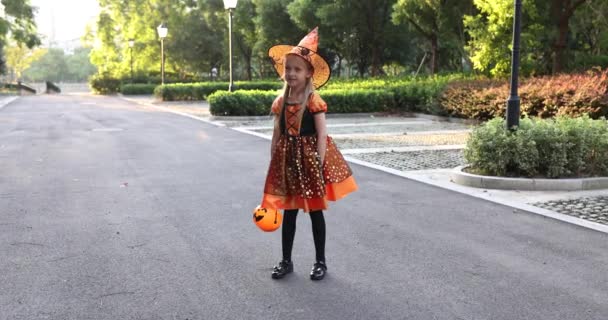 Schattig klein blank meisje met blond haar zeven jaar oud in kostuum van heks met hoed en zwarte oranje jurk vieren Halloween buiten op straat. Vakantie concept. Langzame beweging. — Stockvideo