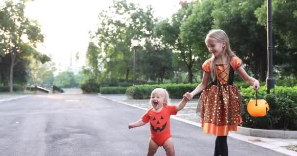 Leuke kleine blanke kinderen met blond haar zeven jaar oud in kostuum van heks met hoed en baby een jaar oud vieren Halloween buiten op straat. Vakantie concept. Langzame beweging. — Stockvideo
